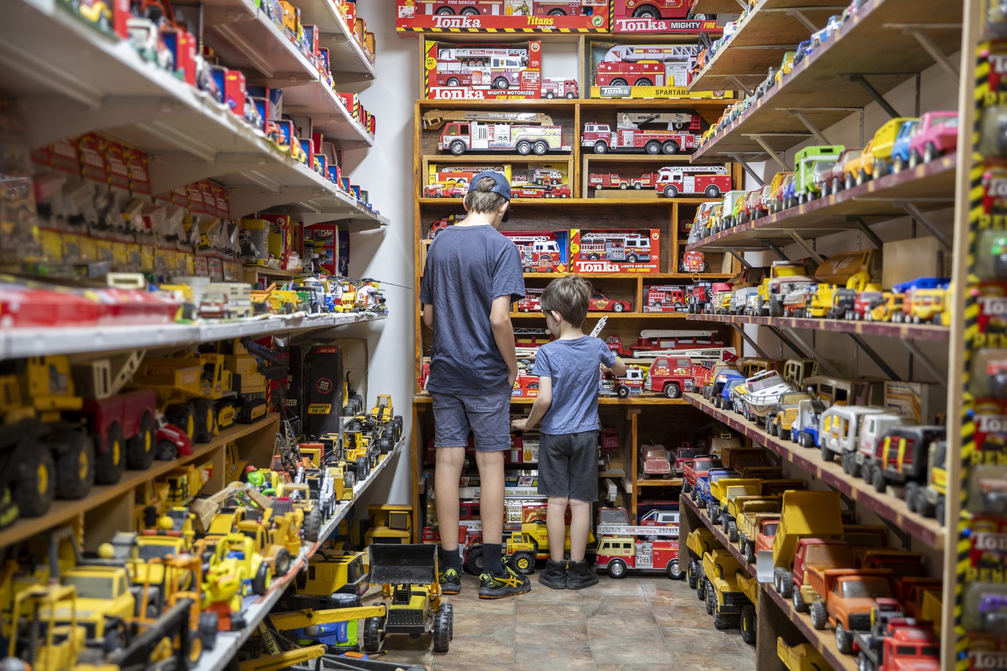 Two children seen from behind, looking up at shelves packed with toy fire engines and trucks